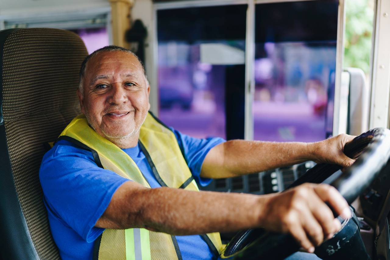 Älterer Mann in Arbeitskleidung mit gelber Warnweste sitzt lächelnd am Steuer eines Busses.