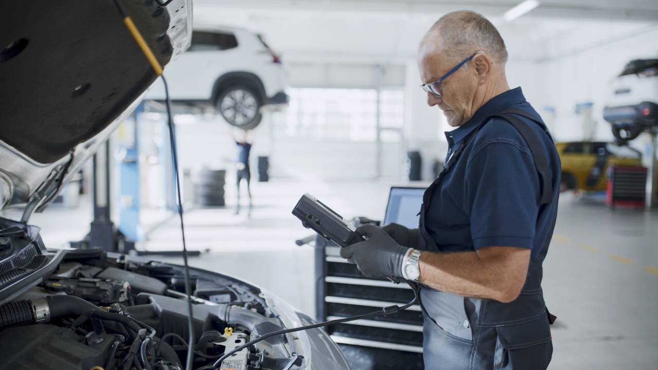 Älterer Mann in Arbeitskleidung arbeitet an einem Auto in einer Kfz-Werkstatt.