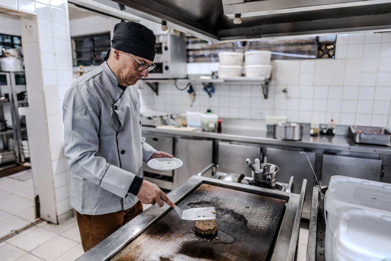 Älterer Koch in typischer Berufskleidung bereitet Speisen auf einer großen Grillplatte in einer Großküche zu.