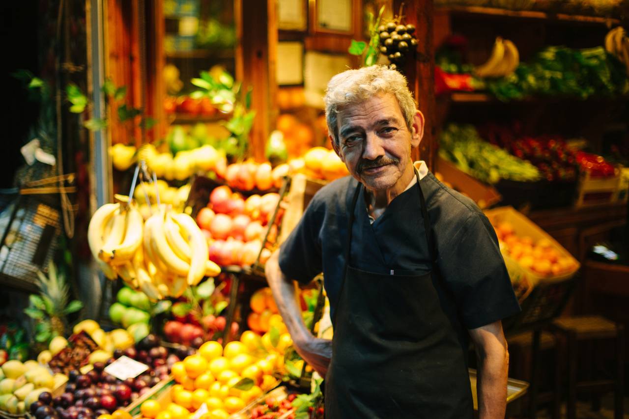 Älterer Mann mit Schürze steht lächelnd vor der farbenfrohen Auslage eines Obst- und Gemüsestandes.