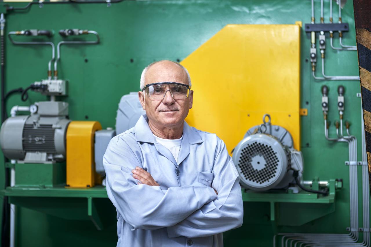 Älterer Mann in hellblauem Arbeitskittel steht mit verschränkten Armen lächelnd vor einer Industriemaschine.