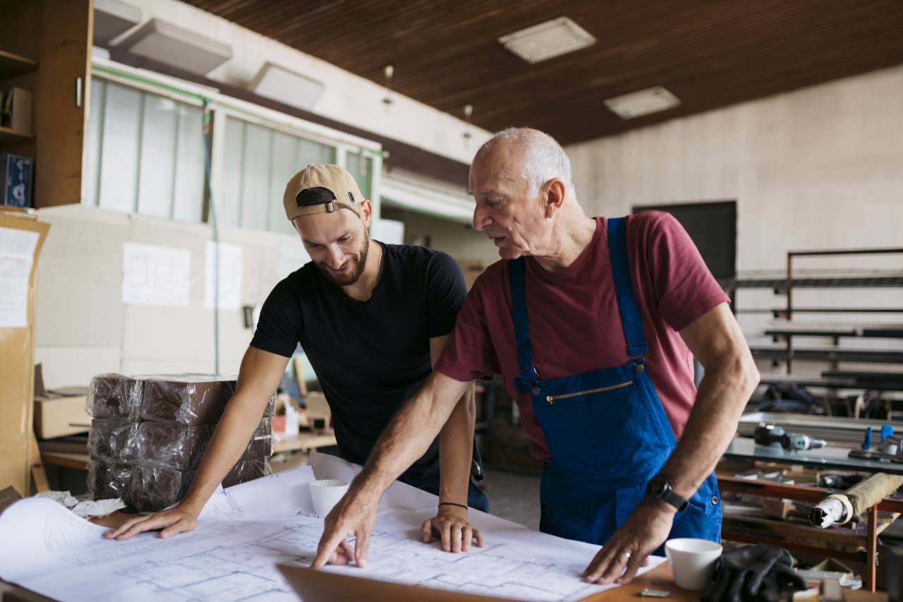 Ein jüngerer und ein älterer Handwerker stehen an einem Tisch in einer Werkstatt und besprechen gemeinsam einen großen Bauplan.