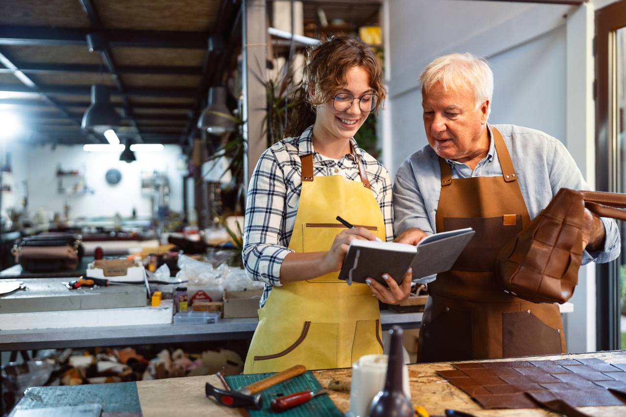 Eine junge Frau in Arbeitskleidung und ein älterer Kollege mit Lederschürzen stehen in einer Werkstatt und schauen gemeinsam in ein Notizbuch.