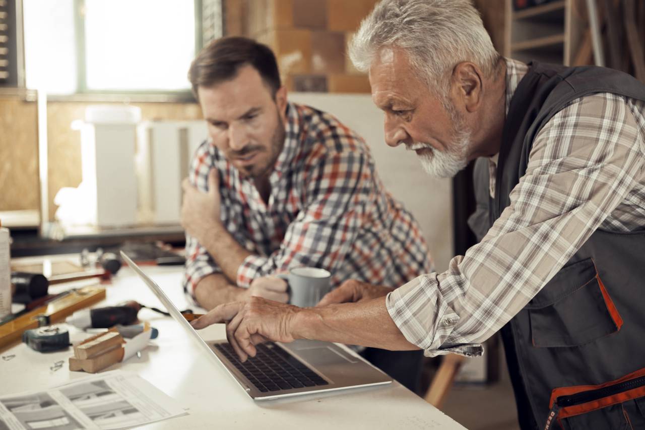 Ein älterer und ein jüngerer Handwerker besprechen gemeinsam einen Bauplan an einem Werkstatttisch.